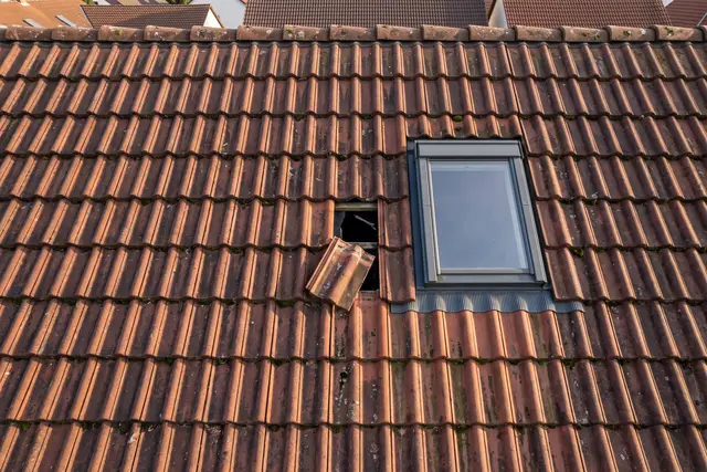 Illustratives Motiv: Nahaufnahme eines Ziegeldachs mit einem gerissenen, verrutschten Ziegel neben einem Dachfenster