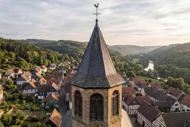 Illustratives Motiv: Kirchturm mit Schieferhelm und Schallöffnungen auf Augenhöhe, darunter ein Fachwerkdorf